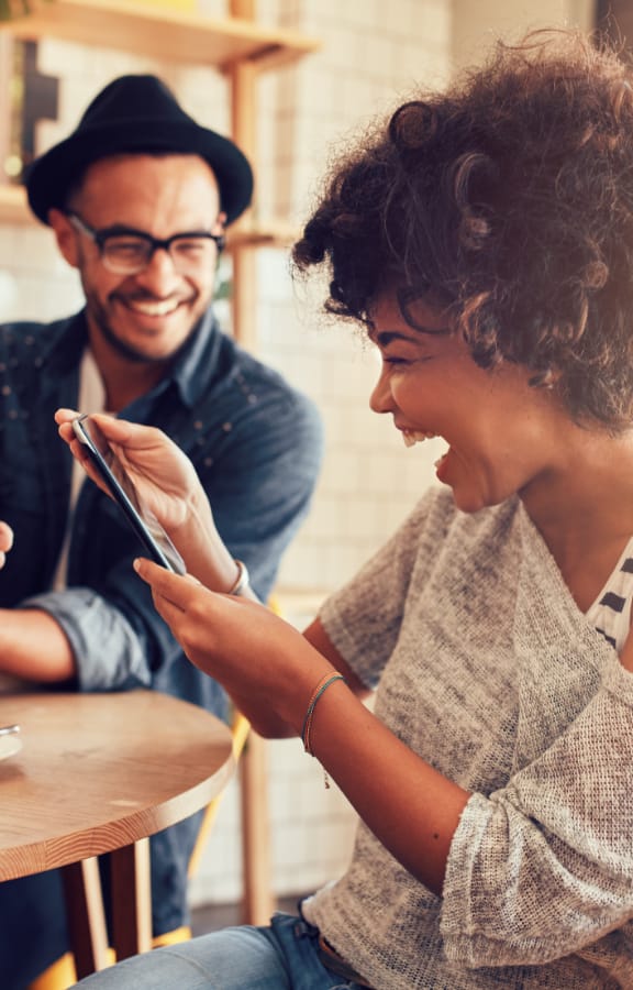 Couple sitting at a table in a coffee shop and laughing at something on the phone they are looking at